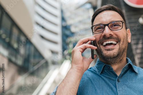 Happy laughing businessman with glasses having telephone call while standing near the office buildings
