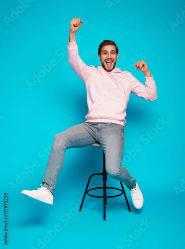 Foto Full length of a cheerful young man sitting on chair and celebrating success isolated over light blue background