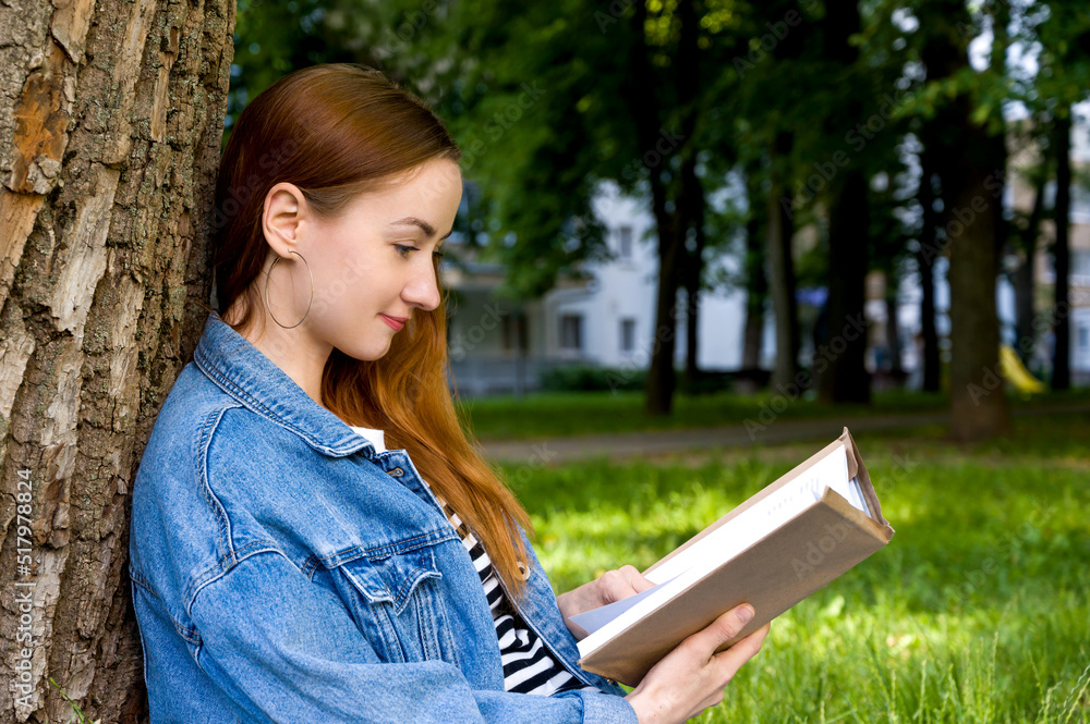 Fototapeta premium Side view portrait of caucasian female student with red hair reading book in the park. Leisure or education concept