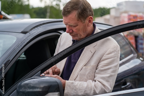 Thoughtful senior business man owner of hardware store with building materials for construction stands with car door open and looks at mobile phone screen