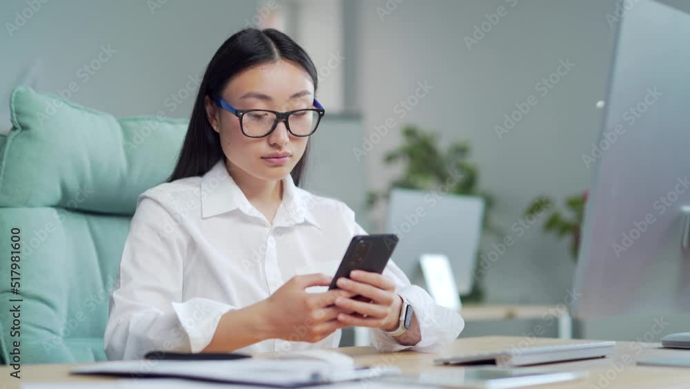 Happy asian business woman employee entrepreneur uses smartphone sitting at workplace in office. young female worker using, browsing, chatting, text message mobile smart phone at computer desk. Smile Happy asian business woman employee entrepreneur uses smartphone sitting at workplace in office. young female worker using, browsing, chatting, text message mobile smart phone at computer desk. Smile