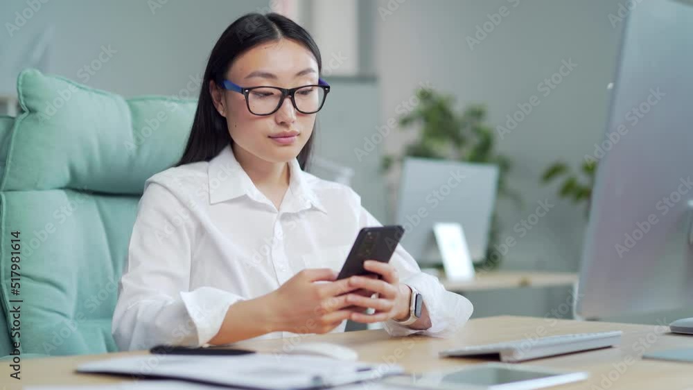 Happy asian business woman employee entrepreneur uses smartphone sitting at workplace in office. young female worker using, browsing, chatting, text message mobile smart phone at computer desk. Smile Happy asian business woman employee entrepreneur uses smartphone sitting at workplace in office. young female worker using, browsing, chatting, text message mobile smart phone at computer desk. Smile