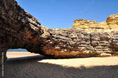 Praia com uma rocha redonda vazia e aberta no meio e com um passagem para fora, oceano e família a passear na areia molhada junto á água do mar