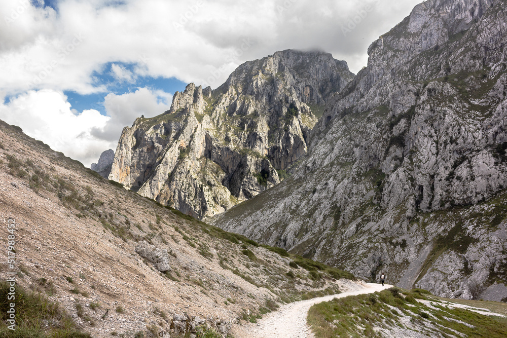 Hiker hiking the Ruta del Cares trail in Picos de Europa National Park, Cantabria, Spain, on a summer day with clouds covering the peaks illuminated by the sun, seeing the path carved in the hillside