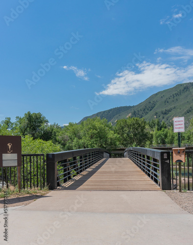Fotografiet Wood Plank and Metal Footbridge Across River with Tree Lined Mountains in Backgr
