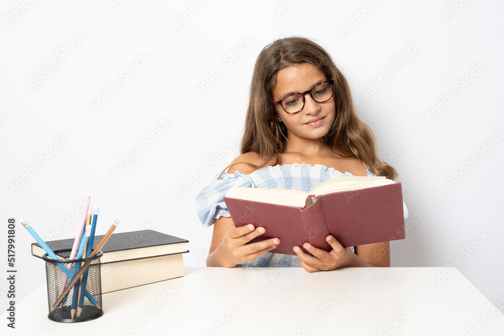 Portrait of positive school girl sitting behind desk reading a book isolated on white background Portrait of positive school girl sitting behind desk reading a book isolated on white background