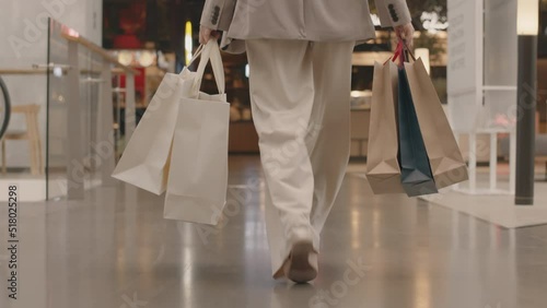 Low section rear view tracking slowmo of unrecognizable young woman in smart casualwear carrying many paper shopping bags walking confidently towards food court in modern shopping center