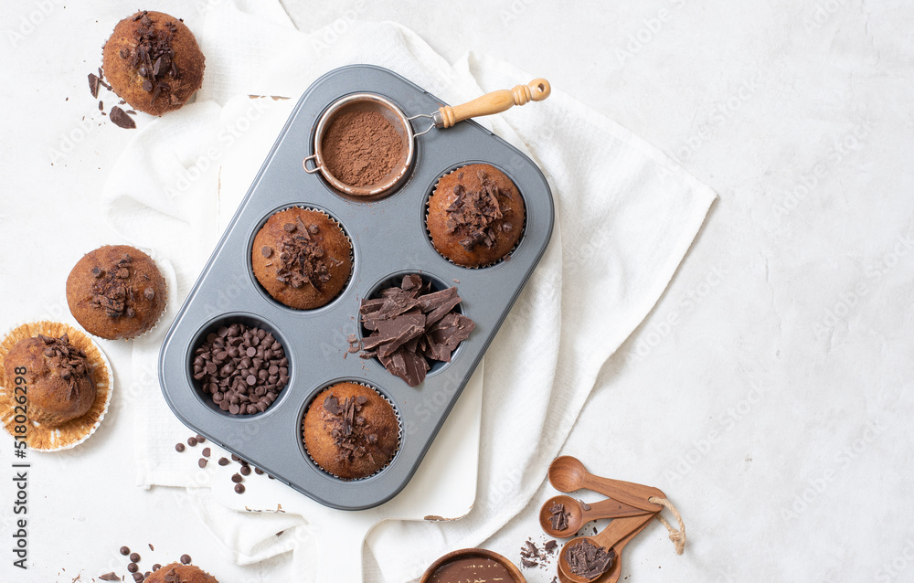 Top view of chocolate muffins flat lay in baking tray with slides of chocolate, chocolate chip, cocoa powder and chocolate sauce on white cutting board and white cloth with copy space Top view of chocolate muffins flat lay in baking tray with slides of chocolate, chocolate chip, cocoa powder and chocolate sauce on white cutting board and white cloth with copy space