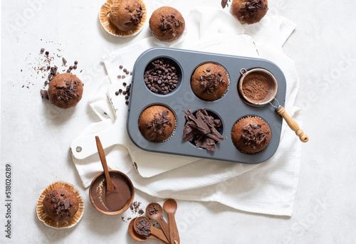 Top view of chocolate muffins flat lay in baking tray with slides of chocolate, chocolate chip, cocoa powder and chocolate sauce on white cutting board and white cloth