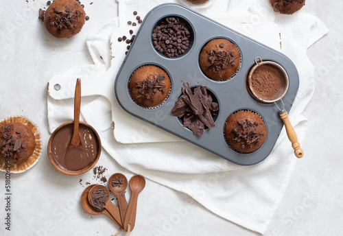 Top view of chocolate muffins flat lay in baking tray with slides of chocolate, chocolate chip, cocoa powder and chocolate sauce on white cutting board and white cloth