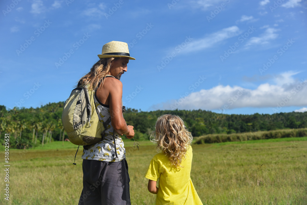 A father and son with long curly hair are walking outdoors. Family trip, hike, trip, vacation, weekend, summer vibe. Green grass and blue sky in the background, lifestyle, back view