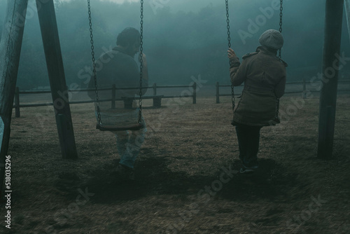 Concept of afterlife and memory for parents or lost love. Back view of woman sitting on a swing with ghost man near her outdoor in the park with fog. . Dead friend or husband concept. Life and death photo