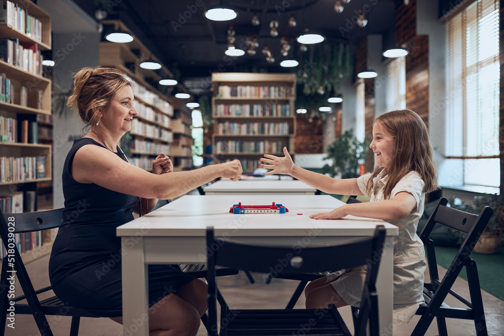 Fototapeta premium Teacher playing with schoolgirl rock, paper, scissors game in afterschool club. Child playing in school library after classes at primary school. Back to school