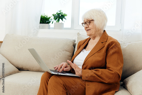 a joyful elderly woman is mastering new technologies using the Internet on a laptop while sitting on the couch at home