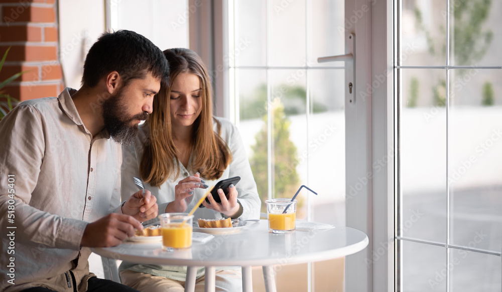 Portrait of beautiful young couple having a break time in cafe, he is showing her something funny in his mobile phone, enjoying