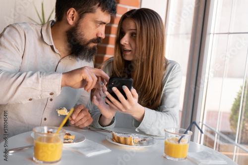 Portrait of happy cute young couple looking at phone and smiling while eating and sitting in a cafe. Joyful couple using cellphones and laughing while having breakfast in cozy home kitchen