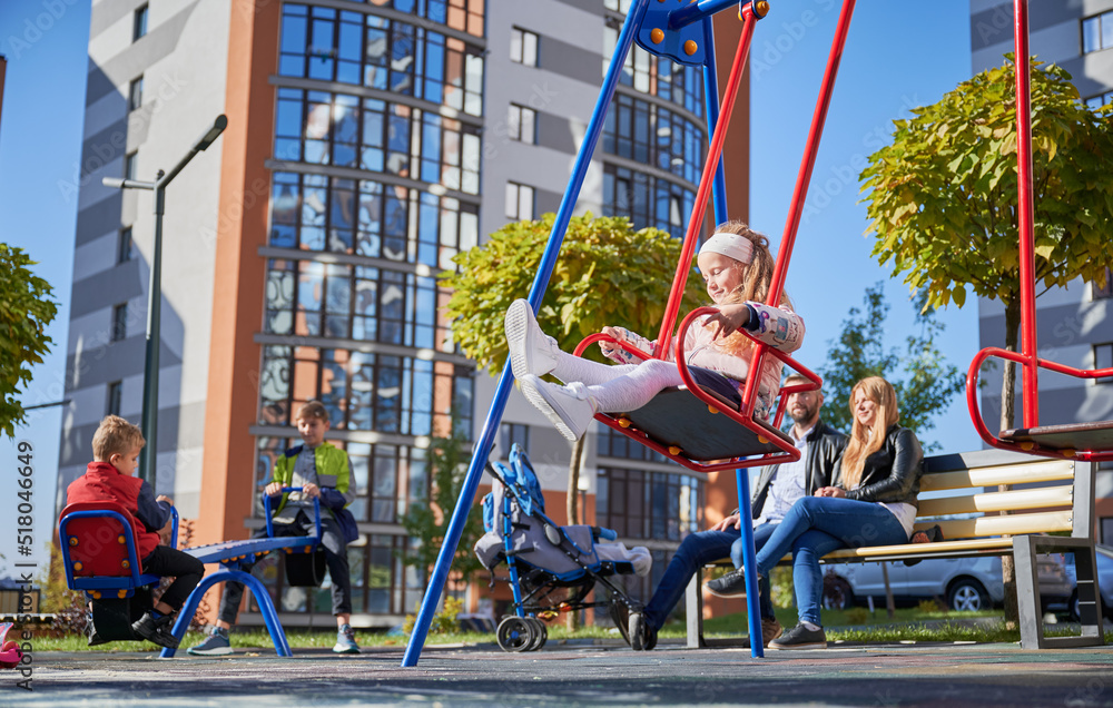 Happy family - father, mother and children having fun together on sunny morning. Parents sitting on bench while kids playing on playground. Modern residential buildings on background.