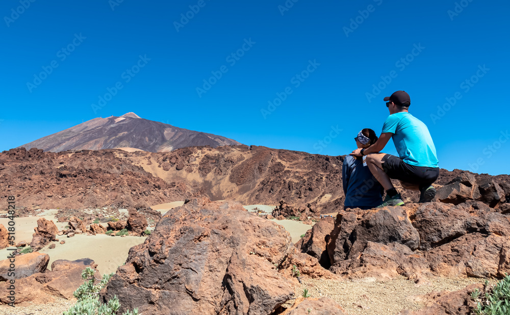 Rear view of hiking couple sitting on lava rock. Scenic view on moon landscape Minas de San Jose Sur near volcano Pico del Teide, Mount El Teide National Park, Tenerife, Canary Islands, Spain, Europe