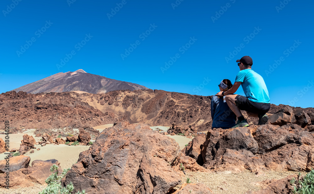 Rear view of hiking couple sitting on lava rock. Scenic view on moon landscape Minas de San Jose Sur near volcano Pico del Teide, Mount El Teide National Park, Tenerife, Canary Islands, Spain, Europe Rear view of hiking couple sitting on lava rock. Scenic view on moon landscape Minas de San Jose Sur near volcano Pico del Teide, Mount El Teide National Park, Tenerife, Canary Islands, Spain, Europe