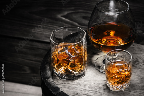 Cognac, whiskey, brandy poured into glasses stand on wooden barrel with alcoholic beverage whiskey close-up. Alcoholic beverages in glasses with ice on black background.