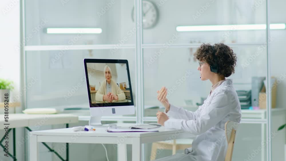 Female doctor in headset sitting at workplace in clinic and having talk with eldelry woman via online video call on computer while consulting remotely during lockdown Female doctor in headset sitting at workplace in clinic and having talk with eldelry woman via online video call on computer while consulting remotely during lockdown