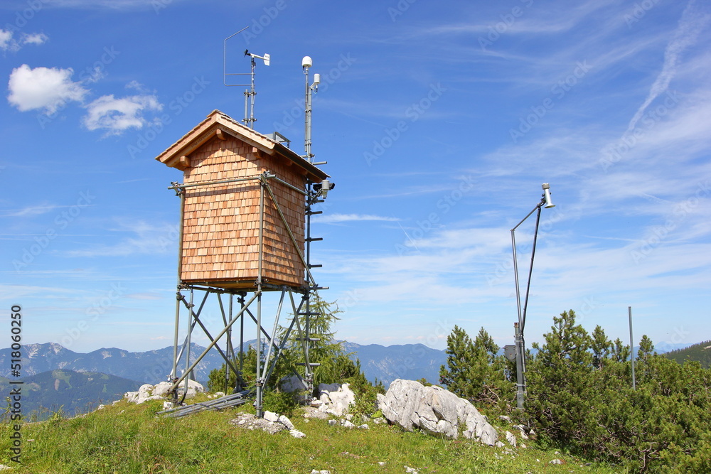 Mountain top meteorological (weather) station on a small cabin with a blue sky, mountains, and scattered clouds