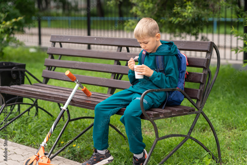 a boy in a blue sweatshirt sits on a bench in the park and eats ice cream from a cup a boy in a blue sweatshirt sits on a bench in the park and eats ice cream from a cup