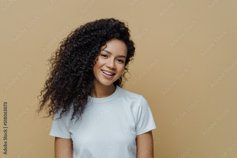 Portrait of beautiful woman with frizzy bushy hair, gentle smile and healthy skin, wears casual white t shirt, enjoys pleasant conversation stands against beige background copy space. People, emotions Portrait of beautiful woman with frizzy bushy hair, gentle smile and healthy skin, wears casual white t shirt, enjoys pleasant conversation stands against beige background copy space. People, emotions