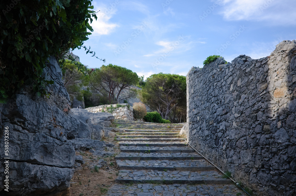 Ancient stone stair leading up between rocks and remains of medieval wall in Porto Venere, Liguria, Italy. Passage between rock and stone wall leading to castle in summer day against blue sky