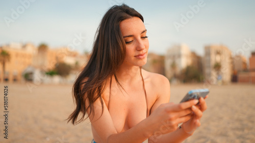 Slim beautiful brown-haired woman with long hair is using mobile phone sitting on the beach. Gorgeous smiling girl scrolling information into smartphone.
