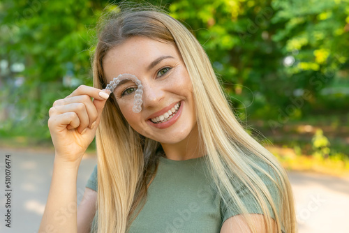 Young beautiful blonde woman holding dental aligner orthodontic to teeth correction with happy face near eyes photo