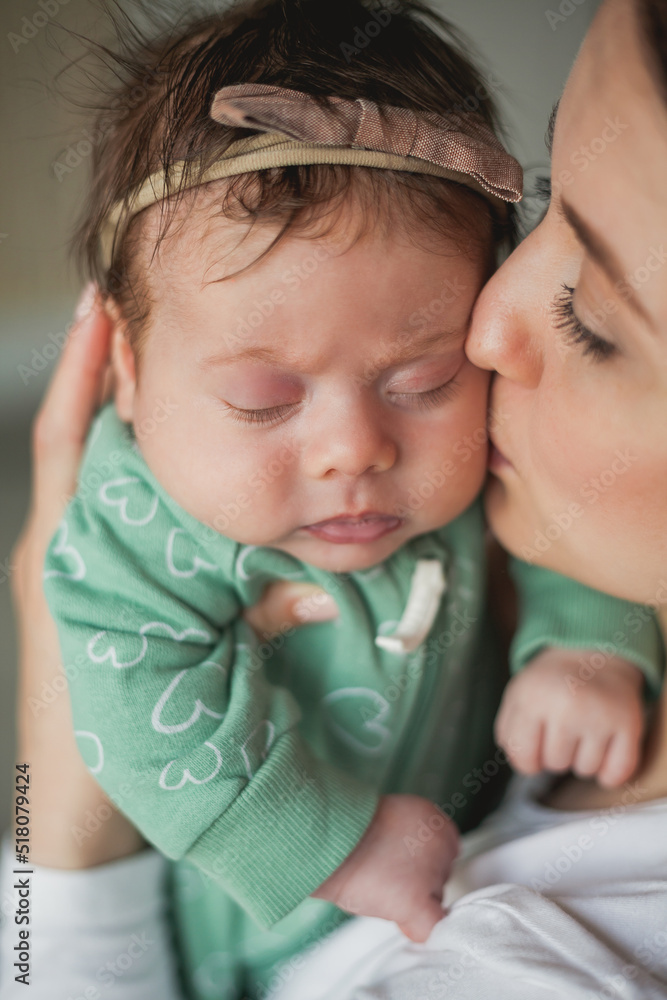 Young beautiful dark-haired mother holds her newborn daughter in her arms in a cozy home. Family portrait. Motherhood. Young woman kisses her 2 month old baby. Young beautiful dark-haired mother holds her newborn daughter in her arms in a cozy home. Family portrait. Motherhood. Young woman kisses her 2 month old baby.