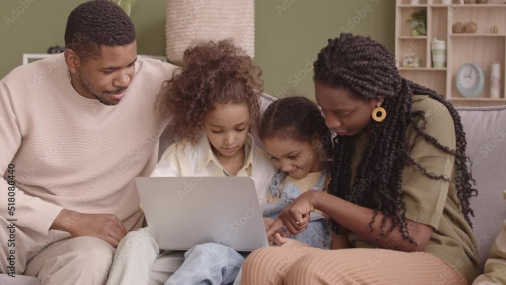 Medium slowmo of happy African American family of four sitting on sofa with laptop, spending weekends together watching films and talking Medium slowmo of happy African American family of four sitting on sofa with laptop, spending weekends together watching films and talking