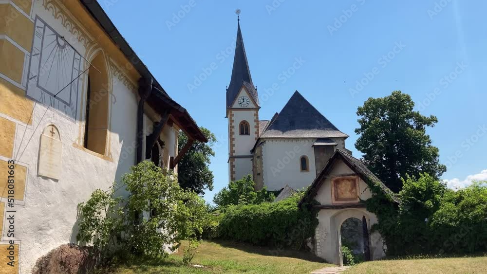 A small decorated church with a small gate and bushes. Sundial on the wall. Clear blue sky. Austrian architecture. A small decorated church with a small gate and bushes. Sundial on the wall. Clear blue sky. Austrian architecture.