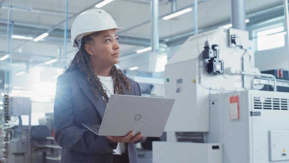 Portrait of a Black Female Engineer in Hard Hat Standing and Using Laptop Computer at Electronic Manufacturing Factory. Technician Working on Daily Tasks and Research and Development Data. Portrait of a Black Female Engineer in Hard Hat Standing and Using Laptop Computer at Electronic Manufacturing Factory. Technician Working on Daily Tasks and Research and Development Data.