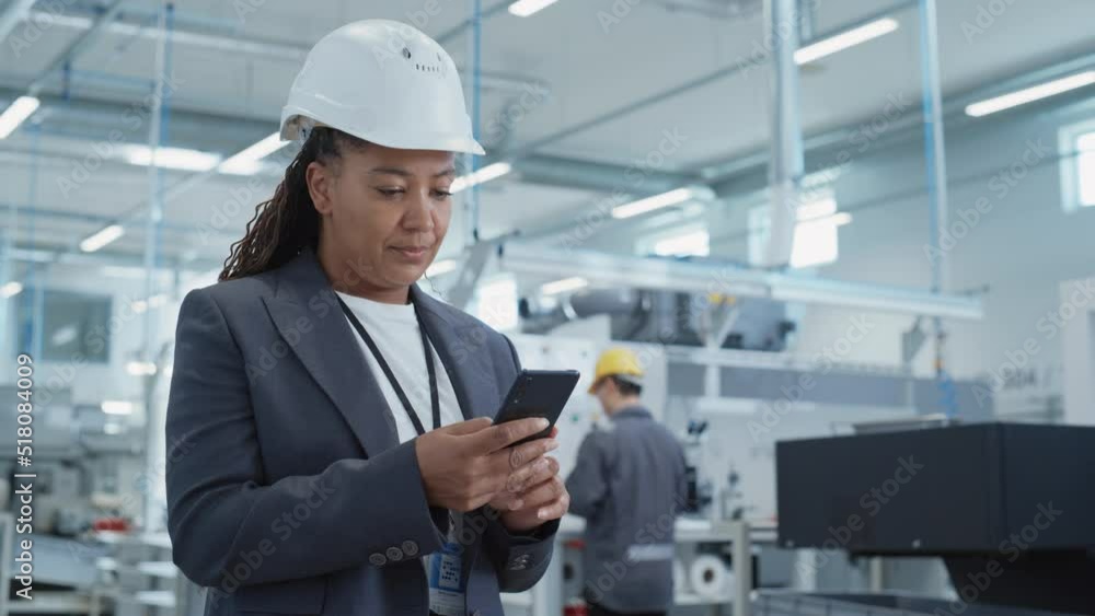 Portrait of a Black Female Engineer in Hard Hat Standing and Using a Smartphone at Electronics Manufacturing Factory. Technician is Writing a Message and Checking Her Schedule. Portrait of a Black Female Engineer in Hard Hat Standing and Using a Smartphone at Electronics Manufacturing Factory. Technician is Writing a Message and Checking Her Schedule.
