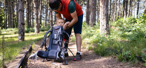 Young Caucasian father puts his little girl in a baby carrier.  They are Going on a hiking adventure  in a nearby forest. It's a lovely sunny summer day 

