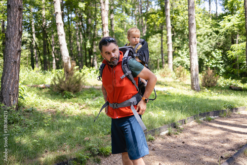 Young Caucasian father is getting ready to go for a walk in the forest with his daughter. He is carrying a child in a baby carrier, wearing a sports clothes. It's a nice sunny day outside © Stock Rocket