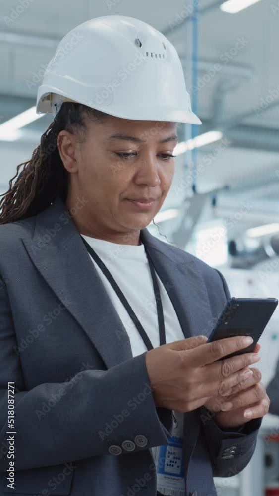 Vertical Screen: Portrait of a Black Female Engineer in Hard Hat Standing and Using a Smartphone at Electronics Manufacturing Factory. Technician is Writing a Message and Checking Her Schedule. Vertical Screen: Portrait of a Black Female Engineer in Hard Hat Standing and Using a Smartphone at Electronics Manufacturing Factory. Technician is Writing a Message and Checking Her Schedule.