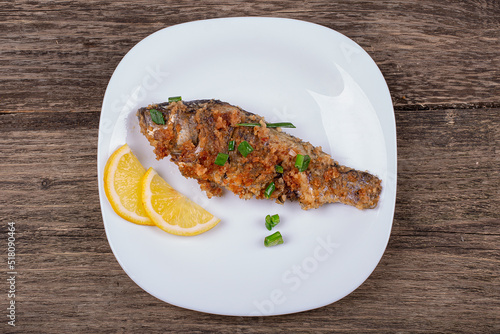Fried breaded fish with green onions and lemon, in a plate on an old wooden background
