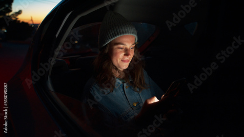 Side night cinematic view of young beautiful woman sit in taxi car back seat near window on road trip, hold, check on mobile phone, use smartphone, scroll social media news, read messages and smile