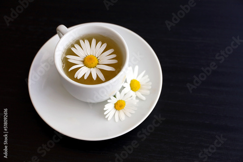 Chamomile tea in a white cup, daisy flowers on saucer. Soothing and healing herbal drink on dark wooden table