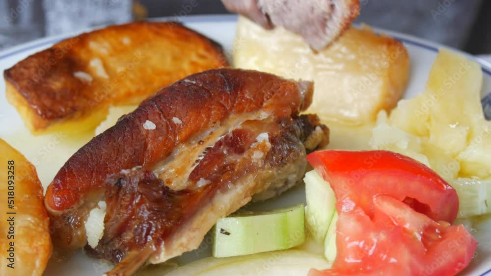 A fork and a knife cut a piece of fried pork meat on a plate in a restaurant. Nearby are boiled potatoes and a salad of tomatoes and cucumbers. Spicy fatty food close up view