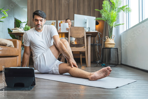 young asian male husband train yoga exercise workout practicing from online lesson at living room with wife working at behind,working from home stay home state order lifestyle concept photo
