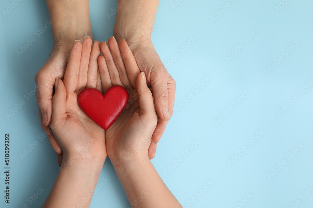 Young and elderly women holding red heart on light blue background, top view. Space for text