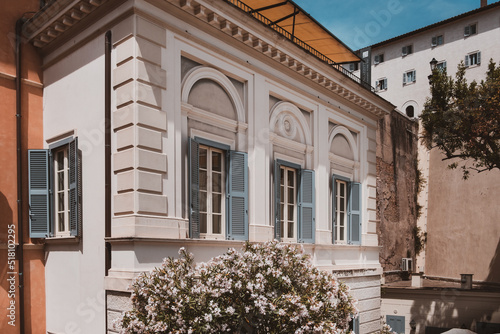 View of corner of an impressive renovated ancient white building with blue window shutters and with blooming green bushes in the foreground