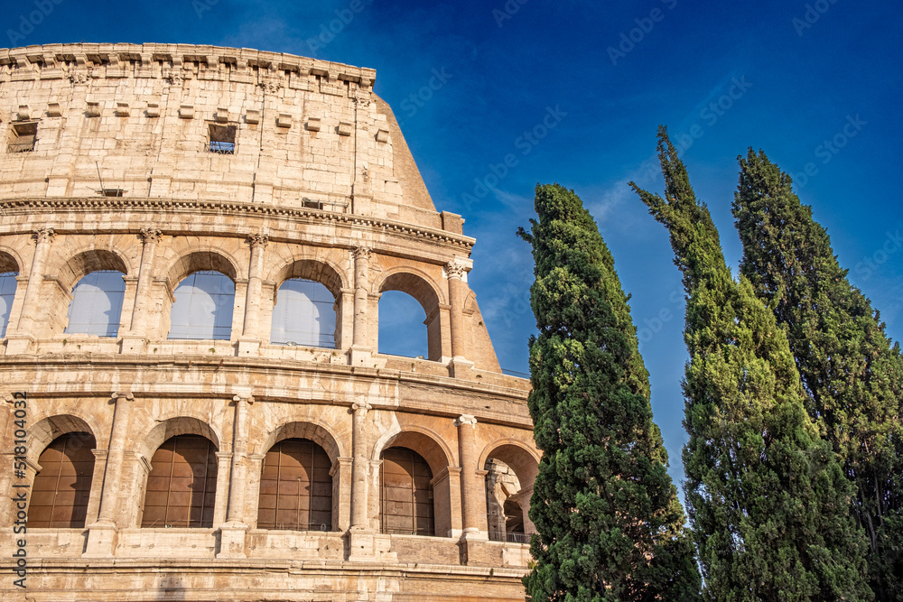Impressive exterior view of Colosseum in Rome with blue sky and clouds in background and with some cypresses in the foreground.