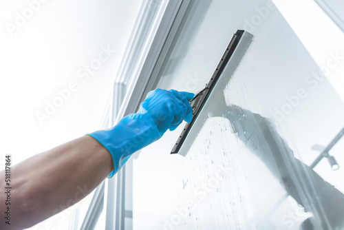 Fototapeta A man wearing light blue rubber gloves cleans the surface of an interior office window with a glass wiper or squeegee