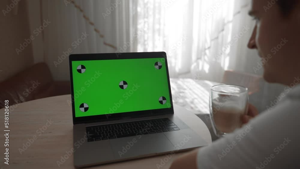 Man drinks cappuccino watching educational lecture on laptop at home. Modern electronic device with green chromakey screen stands on table closeup Man drinks cappuccino watching educational lecture on laptop at home. Modern electronic device with green chromakey screen stands on table closeup