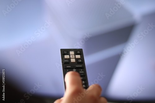 Hand with a remote control, in the background is a TV with a list of channels.
